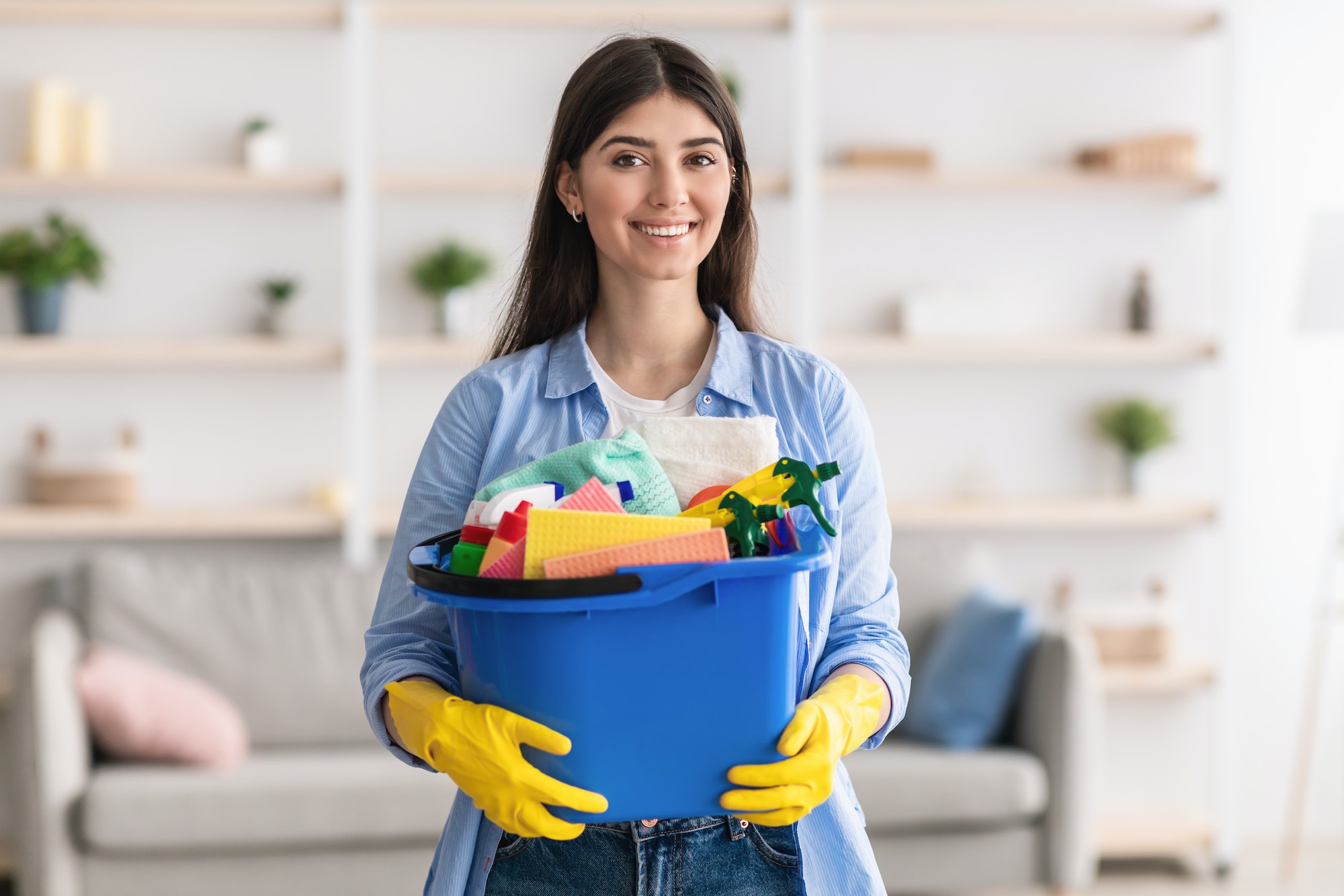 Cheerful young housewife holding bucket with cleaning supplies tools